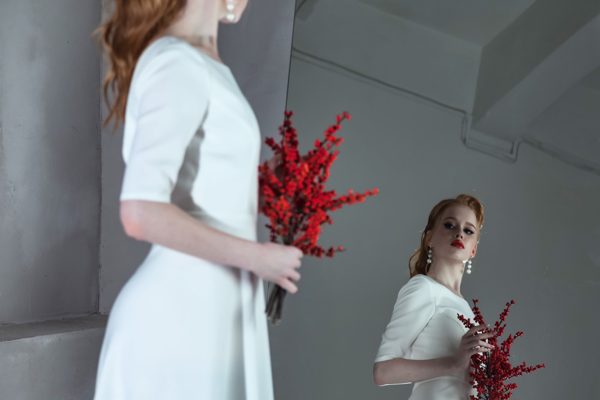 Stylish bride lady with red hair posing at mirror looking reflection. Portrait of fashionable irish woman in white wedding dress with rowan berries bouquet. Fashion style beauty concept. Copy ad space