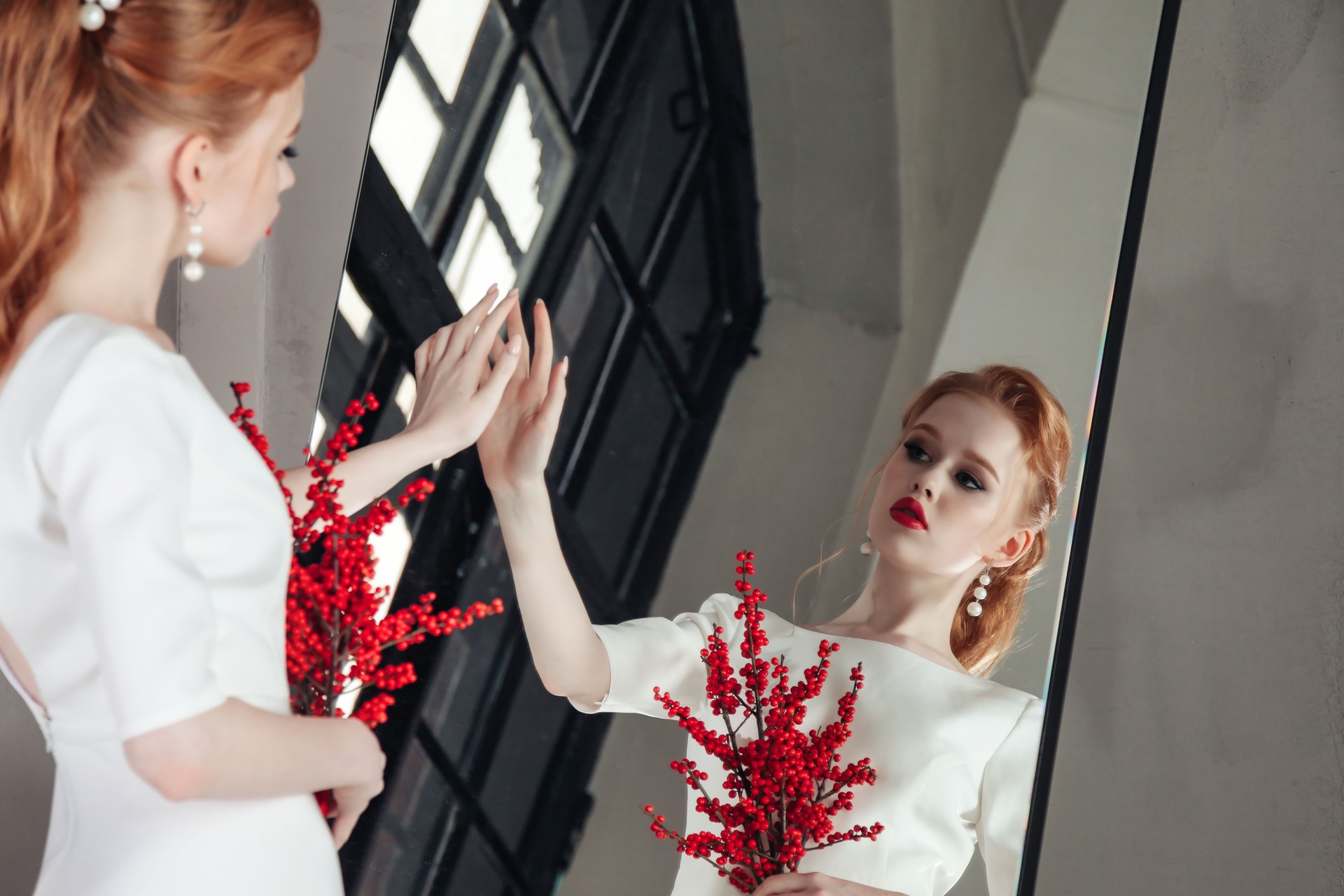 Stylish bride lady looking reflection. Portrait of fashionable irish woman in white wedding dress with red hair posing at mirror with rowan berries bouquet. Fashion style beauty concept. Copy ad space